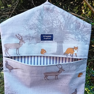 May include: A grey and white fabric hanging organiser with a deer, fox and owl print. The organiser has a blue and white striped pocket and a label that reads "Tiffany & Co. England".
