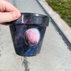 May include: A small, hand-painted flower pot with a cosmic design. The pot has a dark blue and purple background with white speckles, resembling a galaxy. A pink and white planet is painted on the front. The pot is held by a person.