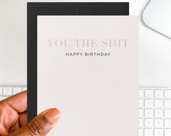 Może przedstawiać: Kartka okolicznościowa z napisem "YOU THE SHIT" i "HAPPY BIRTHDAY" w jasnoróżowej czcionce. Karta jest trzymana przed czarną kopertą. Tło obejmuje białą klawiaturę i komputer.