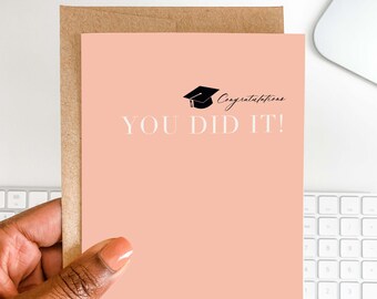 Może przedstawiać: Karta gratulacyjna z okazji ukończenia studiów z brzoskwiniowym tłem i białymi słowami "YOU DID IT!". Czarna czapka absolwenta i słowo "Congratulations" znajdują się nad tekstem. Karta jest trzymana przed brązową kopertą.