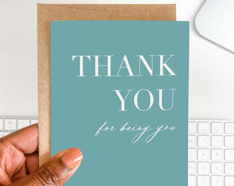 Może przedstawiać: Turkusowa karta z napisem "THANK YOU" dużymi białymi literami i "for being you" kursywą. Karta znajduje się w brązowej kopercie, trzymanej w dłoni.
