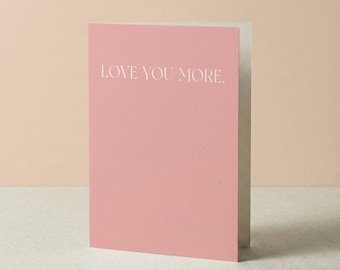 Może przedstawiać: Prostokątna kartka okolicznościowa z jednolitą, pudrowo-różową okładką. Napis LOVE YOU MORE. jest wydrukowany czystą, białą czcionką bezszeryfową w górnej części kartki. Kartka jest lekko uchylona, ukazując białe wnętrze.