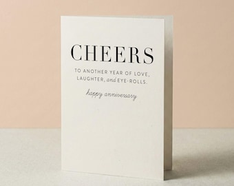 Może przedstawiać: Kremowa kartka okolicznościowa z czarnym napisem CHEERS TO ANOTHER YEAR OF LOVE, LAUGHTER, and EYE-ROLLS. Poniżej znajduje się napis happy anniversary wykonany kursywą. Kartka stoi w pozycji pionowej.