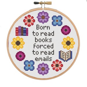 以下が含まれることがあります： 「Born to read books forced to read emails」の文字が入ったクロスステッチ刺繍枠。カラフルな花、本、開いた本がデザインされています。刺繍は木製の枠にセットされています。