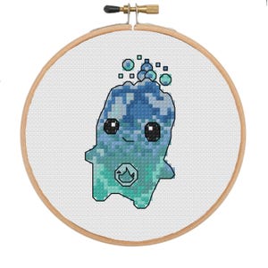 May include: A cross-stitch embroidery project in a round wooden hoop featuring a pixelated blue and teal creature with large black eyes and a small flame symbol on its chest, surrounded by floating bubbles on a white fabric background.