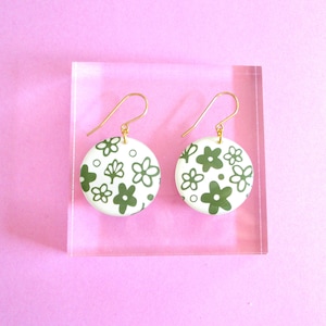 May include: A pair of white, round earrings with a green floral pattern and gold-colored hooks. The earrings are displayed on a clear, rectangular stand against a pink background. The floral design includes various flower shapes and leaf details.