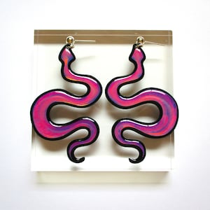 May include: A pair of iridescent pink snake earrings with black outlines. The earrings are made of resin and have a shiny finish.