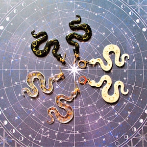 Może przedstawiać: Cztery pary złotych kolczyków w kształcie węża z wzorami niebieskimi. Dwie pary są czarne, jedna para jest biała, a jedna para jest przezroczysta z złotym brokatem.