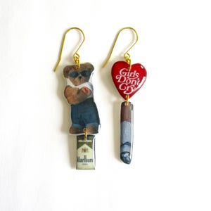 Op de afbeelding: Een paar hangende oorbellen met gouden haakjes. Eén oorbel heeft een rode hartvormige hanger met de tekst "Girls Don't Cry" en een zilveren sigarettenhanger. De andere oorbel heeft een cartoonbeer met een zonnebril en een blauw shirt, en een pakje Marlboro-sigaretten.