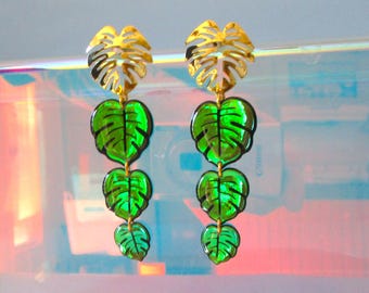 May include: A pair of dangling earrings featuring a gold-colored, stylized leaf design at the top, with three green, leaf-shaped pendants hanging below. The green leaves have black vein detailing. The earrings are set against a blurred, iridescent background.