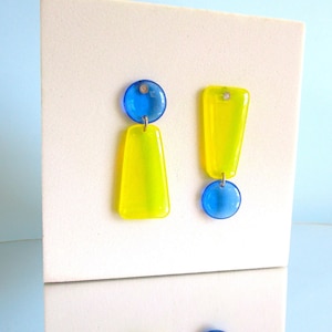 May include: A pair of asymmetrical earrings. One earring features a blue circular top and a yellow, rectangular bottom. The other earring has a yellow, exclamation point-shaped top and a blue circular bottom. The earrings are displayed against a white background.