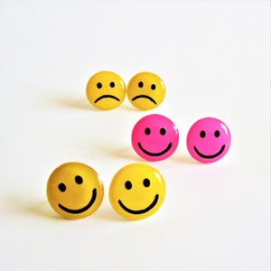 Op de afbeelding: Een verzameling smiley-oorbellen in geel en roze. De oorbellen tonen zowel blije als verdrietige gezichten met zwarte details. De oorbellen zijn rond en hebben een glanzende afwerking.