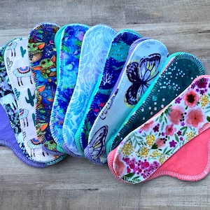 May include: A collection of nine reusable cloth menstrual pads arranged in a fan shape. Each pad features a unique pattern, including floral, butterfly, llama, and character prints, with various colored fabric backings and stitched edges.