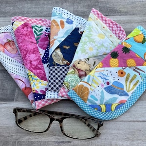 May include: A collection of colorful, quilted fabric items with various patterns, including strawberries, cherries, and floral designs. A pair of eyeglasses with a tortoiseshell frame rests in front of the fabric items.