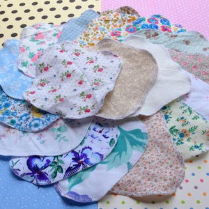May include: A collection of reusable cloth menstrual pads in various floral patterns. The pads feature a variety of colors, including blue, pink, and white, with floral and leaf designs. The edges are neatly stitched, and the pads are arranged in a circular shape.