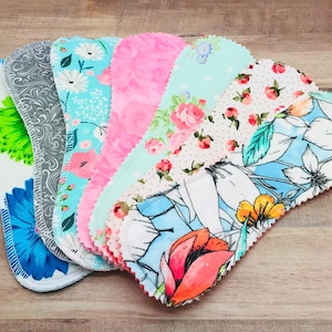 May include: A collection of reusable cloth menstrual pads in various floral patterns. The pads feature designs with flowers in shades of blue, pink, and white. Each pad has a scalloped edge and is made of soft fabric.