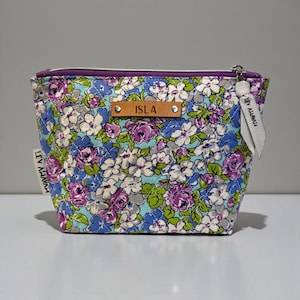 May include: Floral print cosmetic bag with a purple zipper and a leather tag that reads "ISLA". The bag features a colorful pattern of blue, purple, and white flowers. A white tag with the text "LEV AHAVAH" is attached to the side.