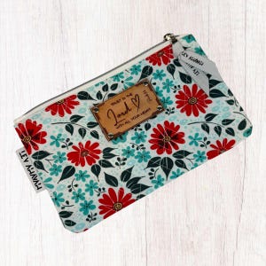 May include: A white zippered pouch with a floral pattern in shades of red, blue, and green. The pouch has a brown leather tag with the text "TRUST IN THE Lord WITH ALL YOUR HEART" and a heart symbol. The pouch also has a white tag with the text "LEV AHAVAH".