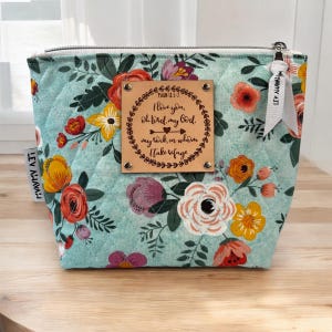 Peut inclure: Une trousse de maquillage matelassée turquoise avec un motif floral rouge, jaune, orange et violet. Un patch en cuir avec un verset biblique est centré sur le devant. La trousse a une fermeture éclair et une étiquette en ruban blanc.