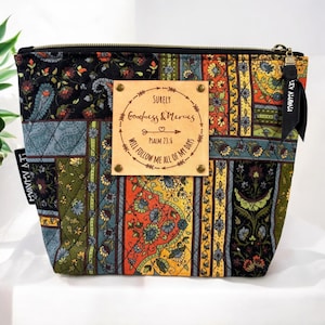 Op de afbeelding: Een zwarte, gewatteerde toilettas met een kleurrijk bloemenpatroon. Een bruin leren patch met de tekst "SURELY Goodness & Mercies PSALM 23:6 WILL FOLLOW ME ALL OF MY DAYS" is bevestigd aan de voorkant van de tas.