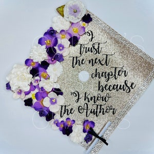 Könnte beinhalten: Eine goldene Glitzer-Abschluss-Kappe, die mit lila und weißen Blumen verziert ist. Auf der Kappe steht der Text "I trust the next chapter because I know the Author".