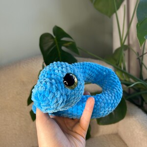May include: A blue crocheted snake toy with a sparkly green eye. The snake is curled into a circle and is being held in a person's hand.
