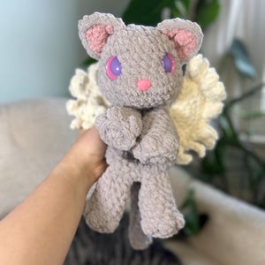 May include: A gray crocheted cat plush toy with pink ears and purple eyes. The cat has white crocheted wings.