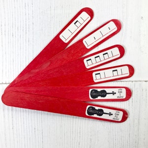 May include: Five red wooden craft sticks with black and white musical notes printed on them. The sticks are arranged in a fan shape. The bottom two sticks have text that reads "oh fiddlesticks!" and "you lose 2 sticks!" and "you lose all sticks!"