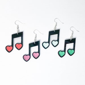 May include: Three pairs of earrings featuring black music note shapes with different colored hearts. One pair has pink hearts, one pair has white hearts, and one pair has green hearts.