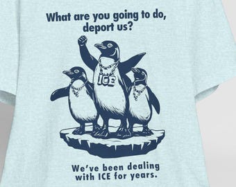 May include: Light blue t-shirt with a graphic of three penguins wearing sunglasses and gold chains. The text "What are you going to do, deport us?" and "We've been dealing with ICE for years." is printed on the shirt.