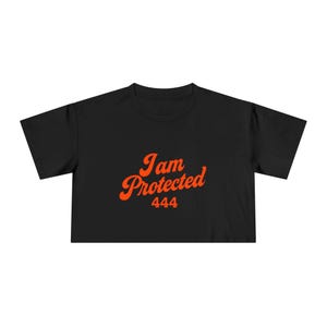 Könnte beinhalten: Schwarzes Crop-Top mit dem orangefarbenen Schriftzug "I am Protected" und der Zahl "444" darunter. Das Shirt hat einen Rundhalsausschnitt und kurze Ärmel.
