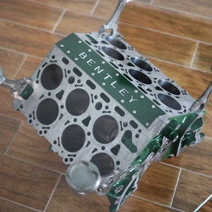 May include: A green and silver engine block coffee table with the word "BENTLEY" engraved on the side. The table has four pistons and connecting rods that act as legs.