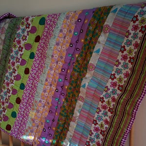 May include: A colorful patchwork quilt with a variety of patterned fabrics, including floral, striped, and polka dot designs. The quilt is draped over a wooden crib, showcasing its vibrant colors and textures.