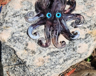Puede incluir: Una figura de pulpo de vidrio con un cuerpo de remolinos en color morado oscuro y negro, acentos blancos translúcidos en las puntas de sus ocho tentáculos curvados y dos ojos de color azul brillante en la parte frontal de su cabeza.