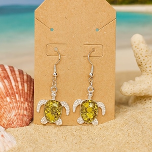 May include: A pair of silver-toned dangle earrings featuring a turtle design. The turtles have a yellow-green glitter-filled body and silver-toned legs and head. The earrings are displayed on a brown earring card, with a beach scene in the background.
