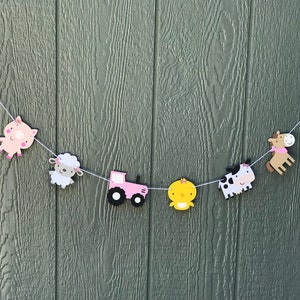 May include: A farm-themed banner with pink barn cutouts, a pig, sheep, tractor, chick, cow, and horse. The banner is strung on white string and is set against a green wooden background.
