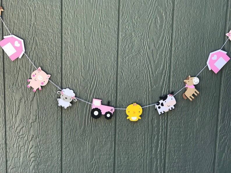 May include: A farm-themed banner with pink barn cutouts, a pig, sheep, tractor, chick, cow, and horse. The banner is strung on white string and is set against a green wooden background.