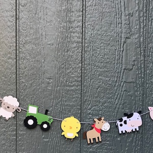 May include: A farm-themed banner with red barns, a green tractor, a yellow chick, a brown horse, a white sheep, a pink pig, and a black and white cow. The banner is strung together with white string.