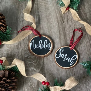 May include: Two round wooden ornaments with black chalkboard paint and white calligraphy. The ornaments are decorated with red and black buffalo plaid ribbon and the names "Nicholas" and "Sarah" are written on them.