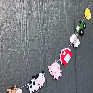May include: A farm-themed paper banner with a red barn, a pig, a cow, a horse, a sheep, a tractor, and a chick. The banner is strung on a white string and is hanging on a gray wooden wall.