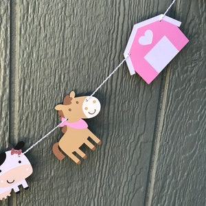 May include: A farm-themed banner with cut-out shapes of a cow, horse, and barn. The cow has a black and white body with a pink bow. The horse is brown with a pink scarf. The barn is pink with a white door and heart. The shapes are strung on a white string.