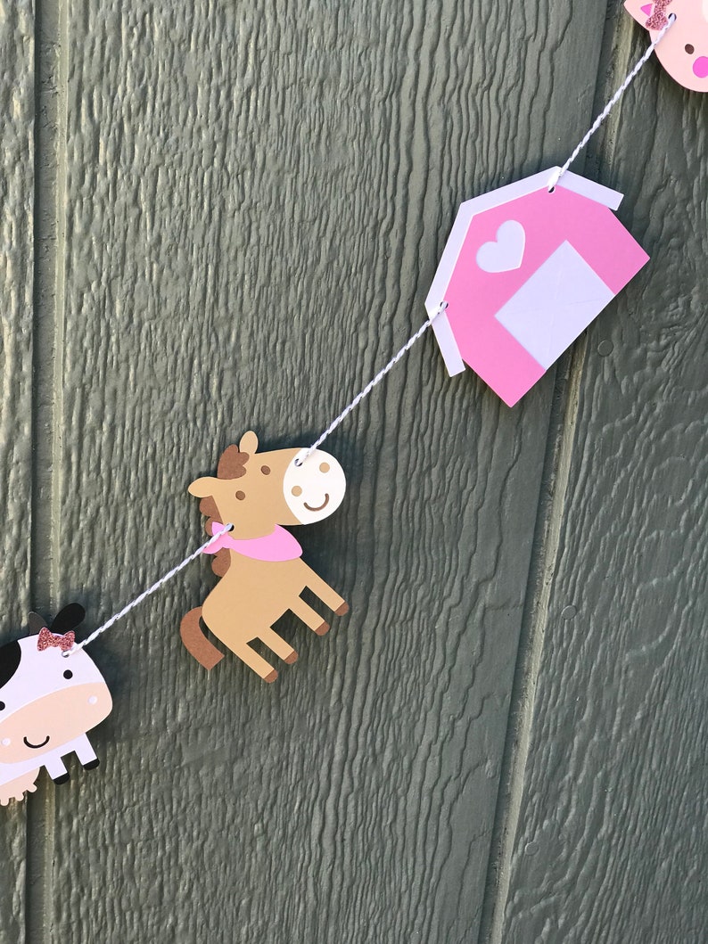 May include: A farm-themed banner with cut-out shapes of a cow, horse, and barn. The cow has a black and white body with a pink bow. The horse is brown with a pink scarf. The barn is pink with a white door and heart. The shapes are strung on a white string.