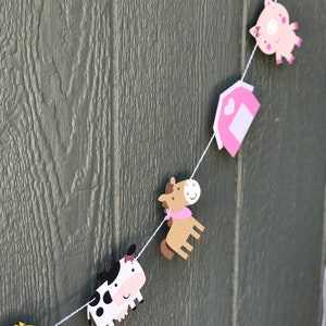 May include: A farm animal-themed banner featuring a cow, horse, pig, and barn cutouts. The cow is black and white, the horse is brown, and the pig is pink. The barn is pink with a white door. The banner is strung on white string.