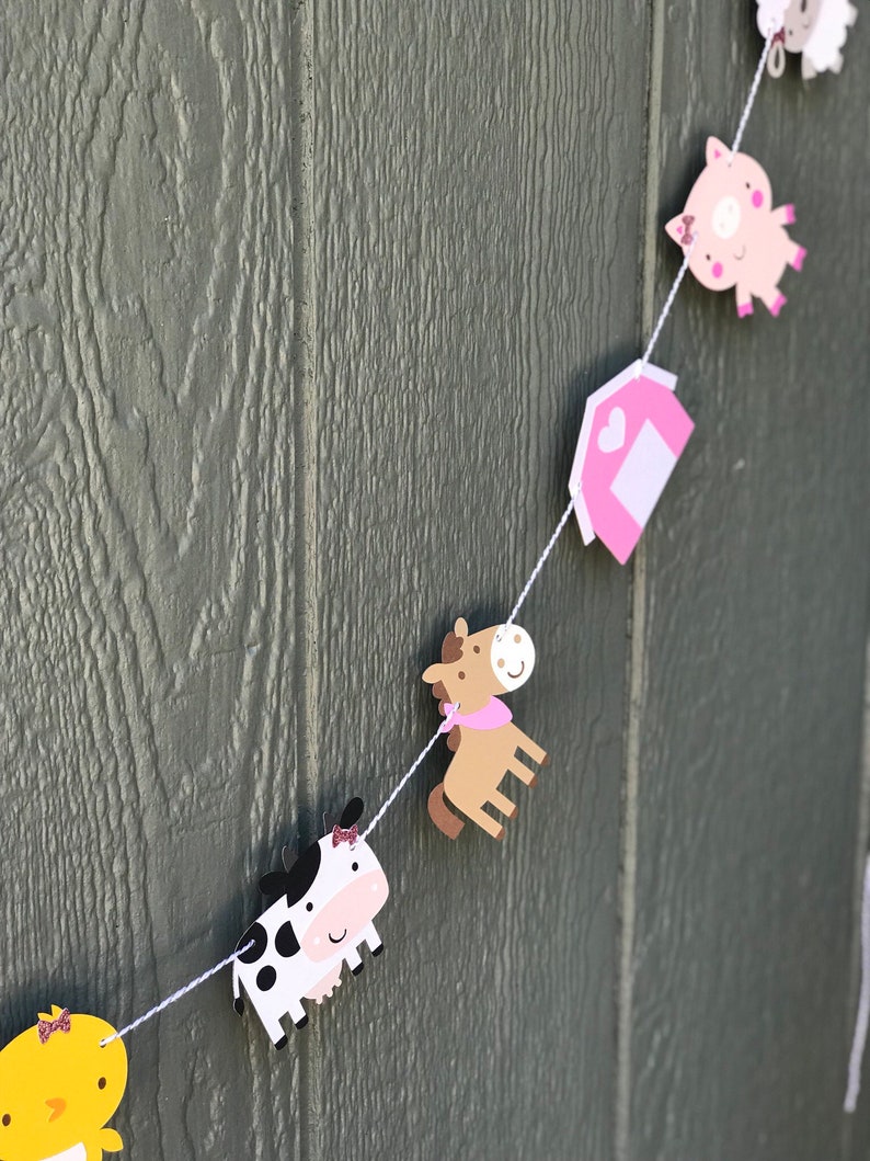 May include: A farm animal-themed banner featuring a cow, horse, pig, and barn cutouts. The cow is black and white, the horse is brown, and the pig is pink. The barn is pink with a white door. The banner is strung on white string.