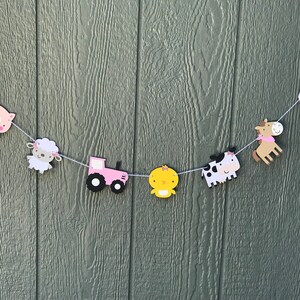 May include: A farm-themed banner with various animal and farm-related cutouts. The banner includes a pink tractor, a yellow chick, a cow, a horse, a pig, a sheep, and a barn. The cutouts are strung together with white string.