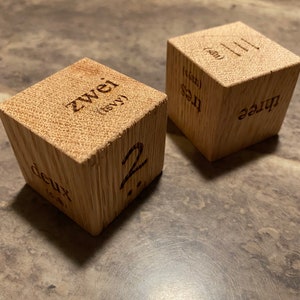 May include: Two wooden blocks with numbers and words in different languages. The first block has the number "2" and the words "deux" and "zwei". The second block has the number "3" and the words "three" and "tres".