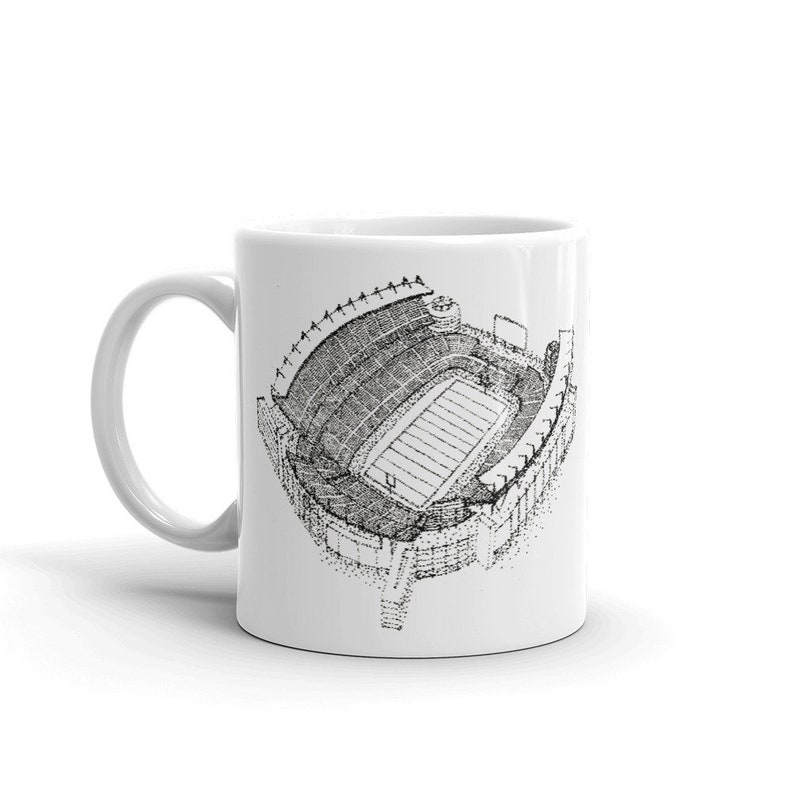 May include: White ceramic coffee mug featuring a black and white illustration of a football stadium.