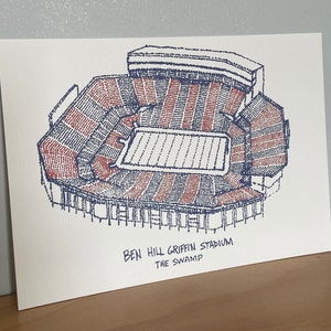 May include: A blue and red line drawing of Ben Hill Griffin Stadium, also known as "The Swamp", a football stadium in Gainesville, Florida.