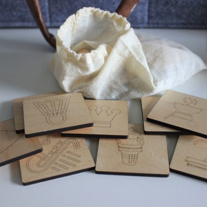 May include: A collection of wooden square coasters with engraved designs, including a badminton shuttlecock, crown, barbecue grill, and ice cream cone. A drawstring bag is visible in the background. The coasters are made of light-colored wood.