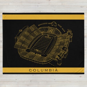 Pode incluir: Cobertor preto e dourado com um contorno detalhado de um estádio de futebol com a palavra "COLUMBIA" na parte inferior.